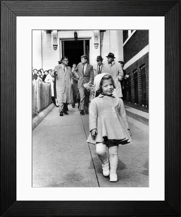 Framed Print : Caroline Kennedy on her third birthday. In the background, her father, President -elect John Kennedy holds her doll as he walks with newsmen and his secret service detail
