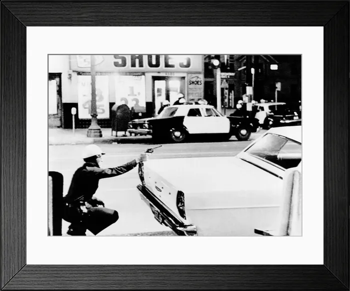 Framed Print : 1965 Watts Riots. A policeman aims his revolver at building where a sniper shot at passing cars. Across the street, fellow officers crouch behind cars as they move in closer