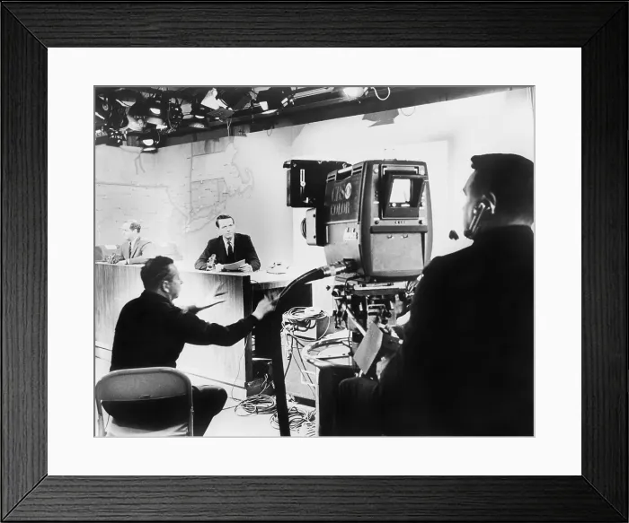 Framed Print : Under hot studio lights, a CBS News anchor performs under the director of his producer (seated between anchor and camera), for a color television camera in 1967 Framed Print : Under hot studio lights, a CBS News anchor performs under the director of his producer (seated between anchor and camera), for a color television camera in 1967