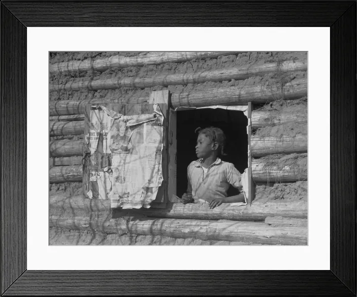 Framed Print : African American girl looking out the window of a plantation log cabin tenants home. Newspaper advertisements on the shutter beckon from the larger world beyond rural poverty