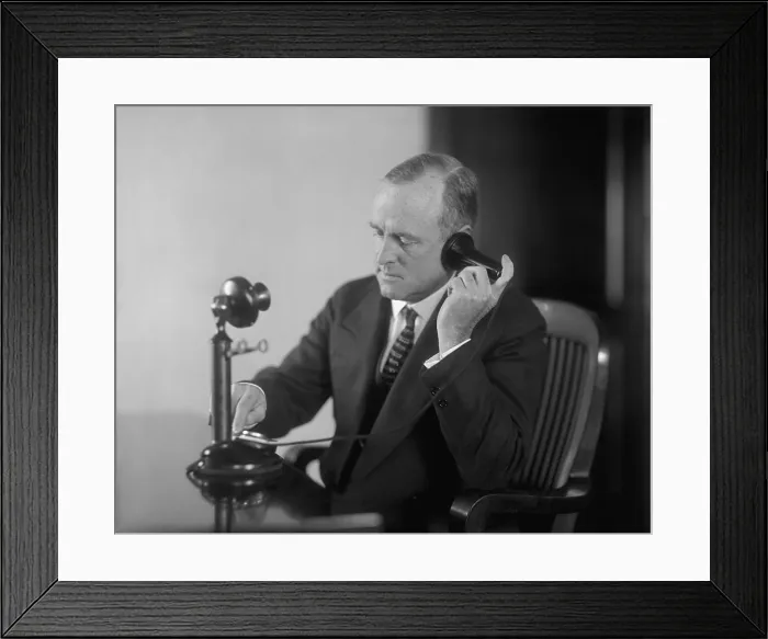 Framed Print : Businessman dialing an early 20th century desk phone with separate ear receiver and mouth transmitter. Ca. 1920