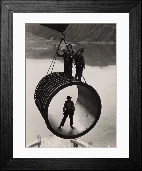 Framed Print : Four workers riding on a large casing section of pipe suspended from cable as it is moved into position during construction (1933-42) of the Grand Coulee Dam, Washington