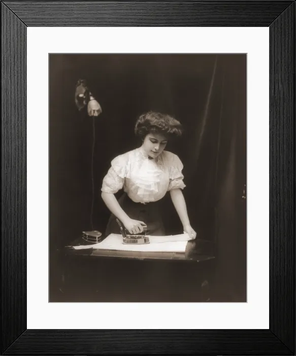 Framed Print : General Electric advertising photo of a woman using an electric iron plugged into light socket, ca. 1908