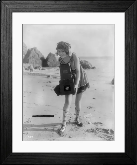 Framed Print : Myrtle Lind, a young woman posing with a Graflex camera on a beach, 1919