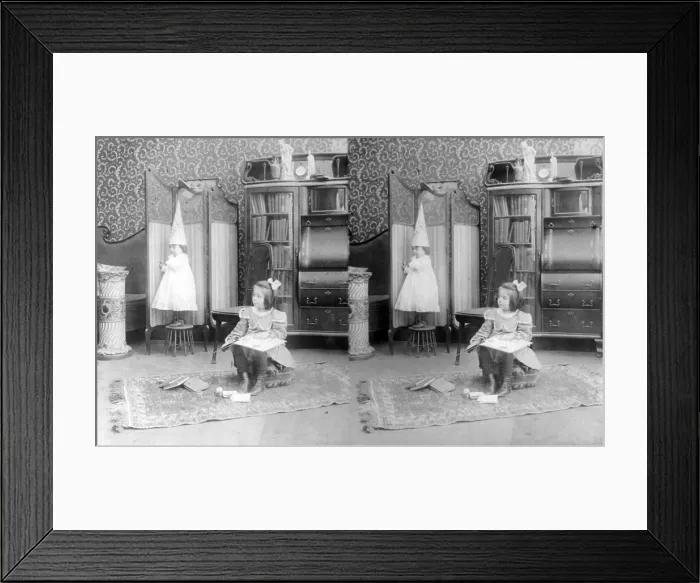 Framed Print : Girl seated in middle of room with books, smaller child standing on stool and wearing dunce cap, stereo photograph, circa 1908 Framed Print : Girl seated in middle of room with books, smaller child standing on stool and wearing dunce cap, stereo photograph, circa 1908