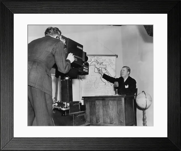 Framed Print : Frazier Hunt, (1885- ) early newscaster, seated at desk, pointing to a wall map of Africa, while being filmed by a cameraman. 1941