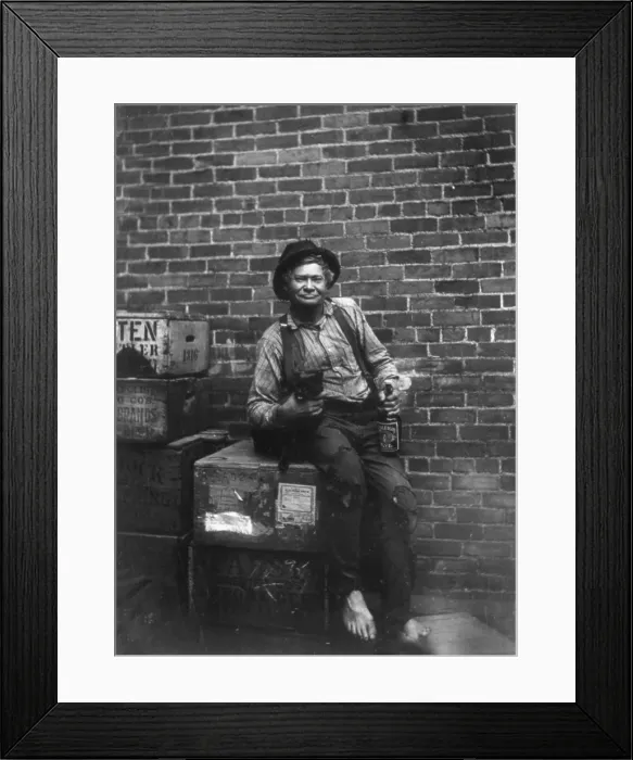 Framed Print : Hobo John Bour, a barefooted, shabbily dressed man, sitting on crates, in front of brick wall, with arm around a cat, holding a bottle of Raleigh Rye whiskey, photograph