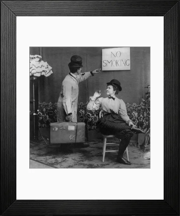 Framed Print : Humorous scene of man pointing to No Smoking sign above woman in derby and bloomers who is smoking cigarette, photograph, 1897