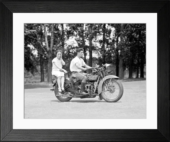 Framed Print : A young man drives a motorcycle while a young woman rides side saddle behind him. ca. 1929-1930