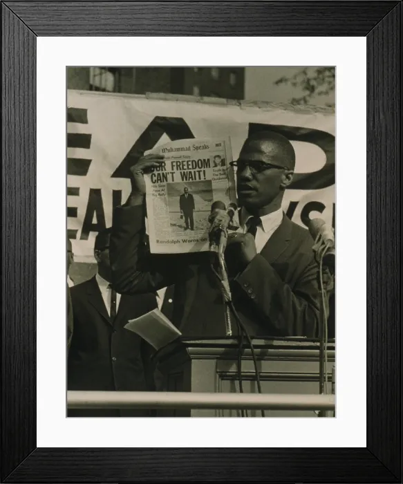 Framed Print : Malcolm X, holding up newspaper with headline Our Freedom Can't Wait, while standing behind podium with microphones addressing a crowd at a Black Muslim rally in New