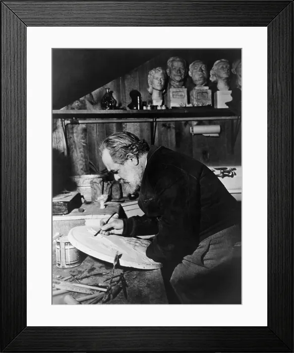 Framed Print : Jo Davidson (1883-1952), Russian American sculptor at work on a marble plaque in his studio, beneath the shelf bearing replicas of his famous portrait busts. Ca. 1940