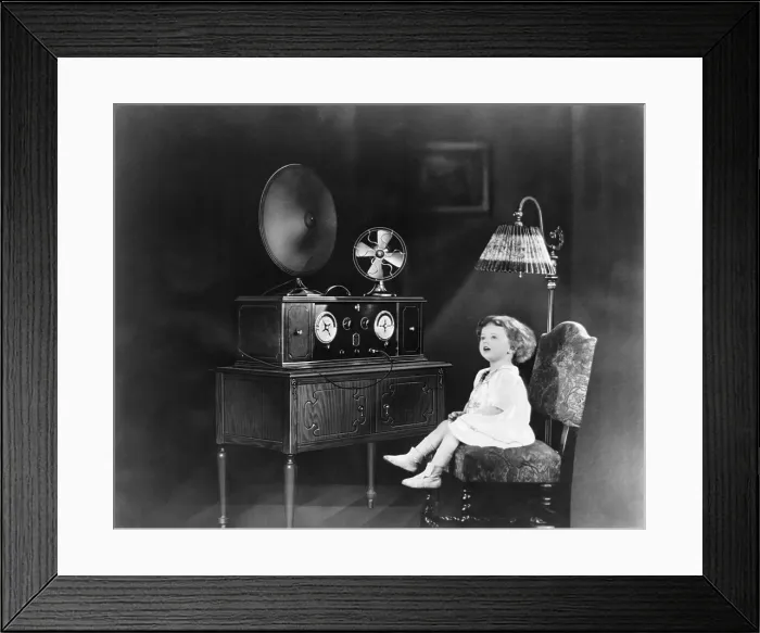 Framed Print : Little girl sitting on chair next to an electric powered radio, with an electric fan and electric light. 1926