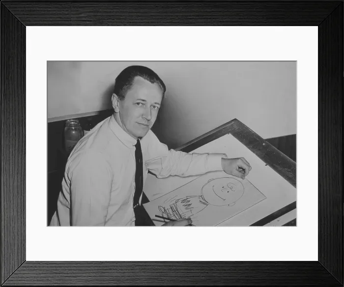 Framed Print : Charles Schulz (1922-2000), seated at drawing table with drawing of Charlie Brown, one of his PEANUTS characters. 1956