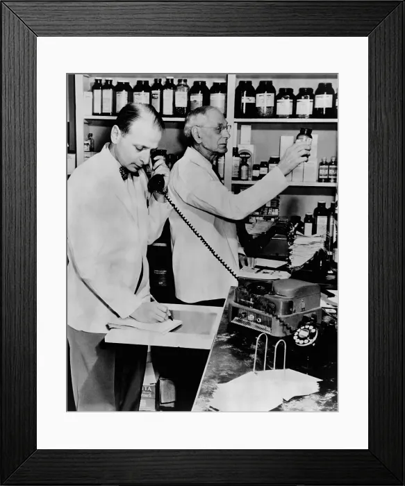 Framed Print : A pharmacist demonstrates the use of an early phone answering machine, a telephone disc prescription recorder, as another fills a prescription. 1952