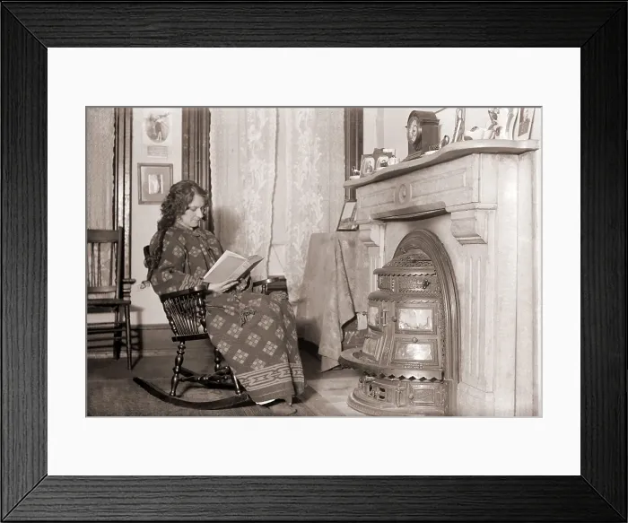 Framed Print : In a heavy robe, a woman reads in front of a coal stove installed in her fireplace