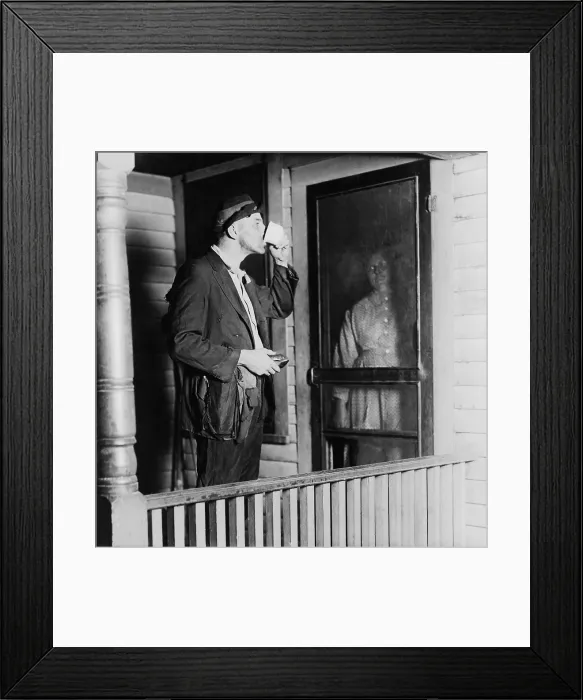 Framed Print : Homeless man in ragged clothes eating a meal standing as his benefactor keeps her distance behind her front door. 1919 photo by N.D. Hillis