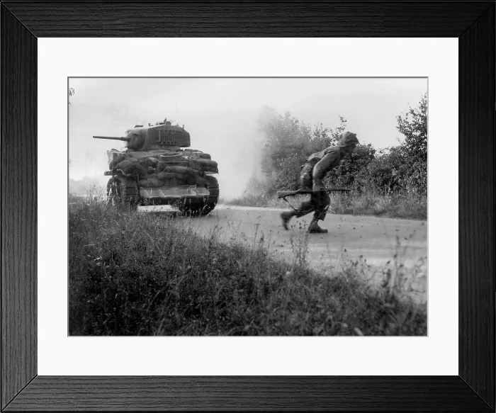 Framed Print : U.S. soldier crosses a road cautiously under the cover of a light tank in the Hedgerows of Normandy. The tank is equipped with the hedgecutter device developed in July 1944