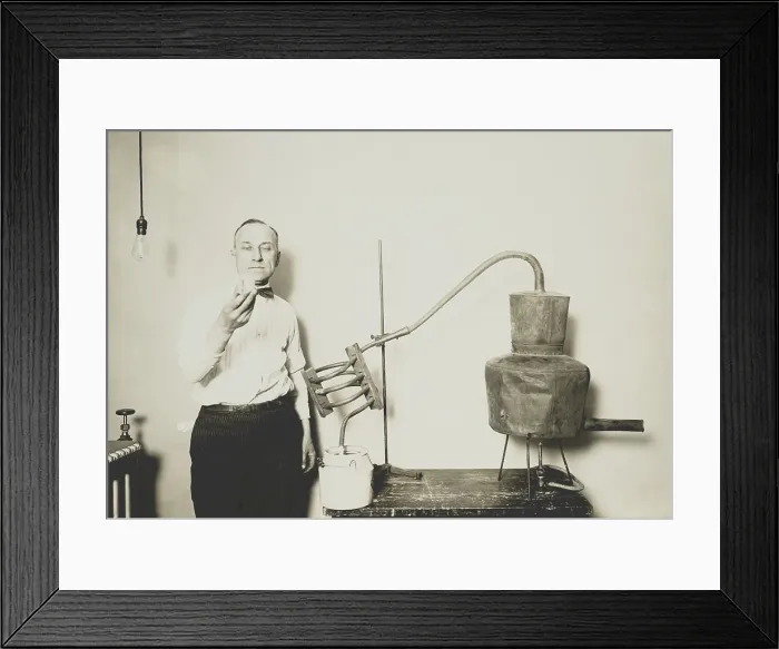 Framed Print : Moonshine still recently confiscated by the Internal Revenue Bureau. An employee holds a glass of alcohol distilled from the apparatus at the Treasury Department, ca. 1921-32