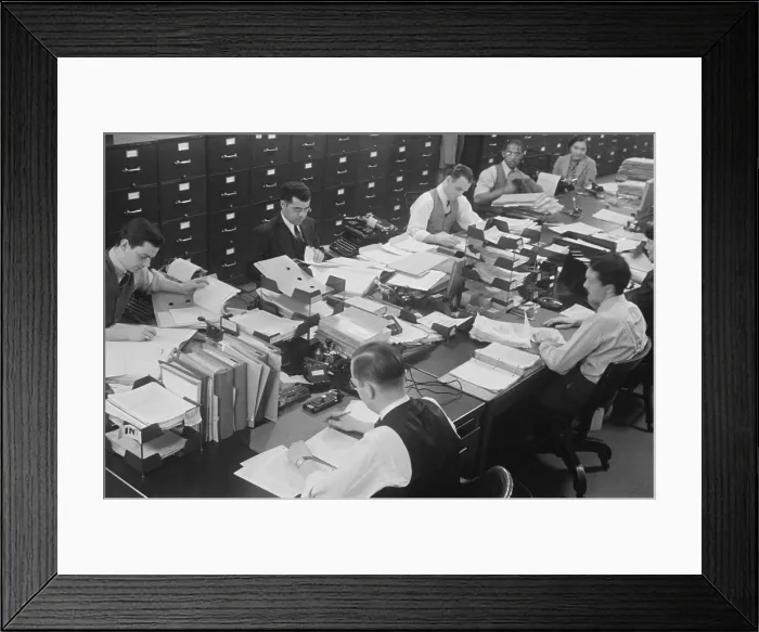Framed Print : Six men and two women at work in a U.S. government office. They share a long table, covered with documents, notebooks, and stacked paper sorters. One women works at a typewriter