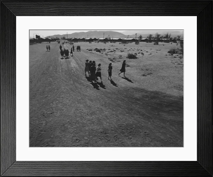 Framed Print : Interned Japanese Americans, mostly women, holding books, walking on a dirt road, within the guarded fences of the Manzanar Relocation Center during World War II