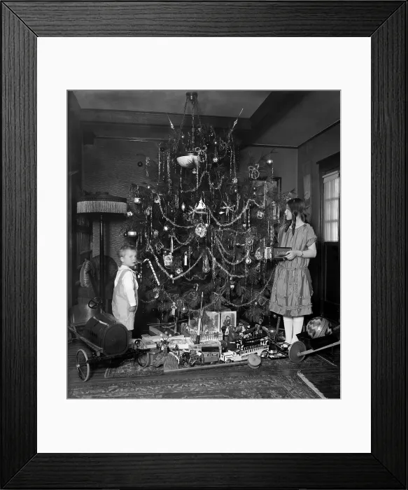 Framed Print : Abundant toys are underneath a traditionally decorated Christmas tree, c. 1920. A boy and girl stand beside holiday tree in their home