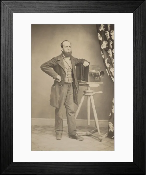 Framed Print : Photographers full length portrait standing next to a box camera resting on a tripod in a photo studio, c. 1866-75. It is possibly of a portrait of Charles L