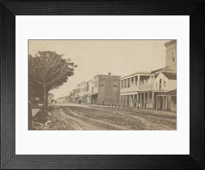 Framed Print : Elizabeth Street in Brownsville, Texas, ca. 1865. Commercial buildings line one side of the dirt road while pedestrians walk on the other side