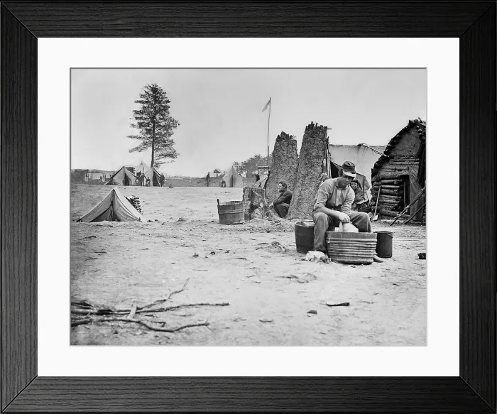 Framed Print : US Civil War, 1861-1865. Camp scene with a soldier does his laundry in a metal tub. Other soldiers are sitting near rough log buildings nearby