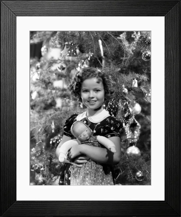 Framed Print : Shirley Temple holding doll in front of Christmas tree, ca. late 1930s