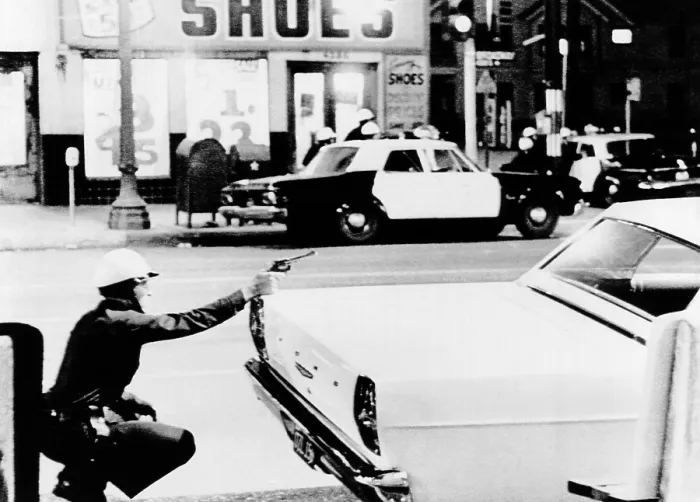 Framed Print : 1965 Watts Riots. A policeman aims his revolver at building where a sniper shot at passing cars. Across the street, fellow officers crouch behind cars as they move in closer