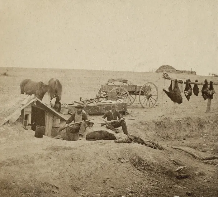 Framed Print : Buffalo hunters dugout home and encampment, ca. 1870. The two hunters pose with their rifles and dog. Unskinned bison meat hangs at right