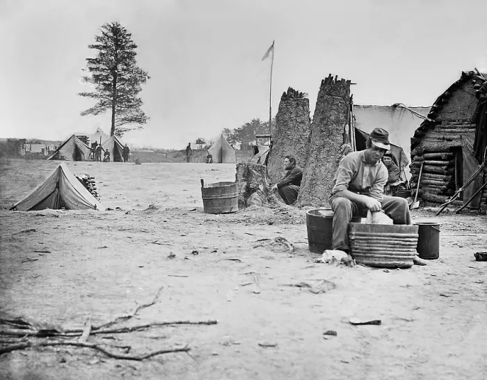 Framed Print : US Civil War, 1861-1865. Camp scene with a soldier does his laundry in a metal tub. Other soldiers are sitting near rough log buildings nearby