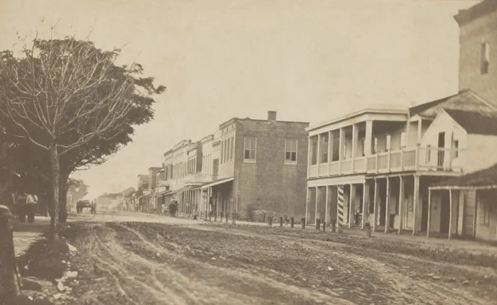 Framed Print : Elizabeth Street in Brownsville, Texas, ca. 1865. Commercial buildings line one side of the dirt road while pedestrians walk on the other side
