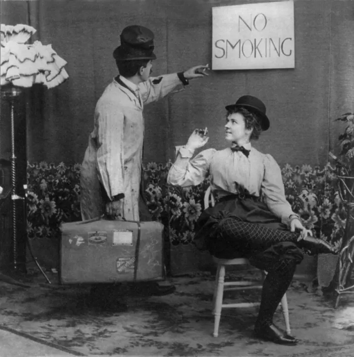 Framed Print : Humorous scene of man pointing to No Smoking sign above woman in derby and bloomers who is smoking cigarette, photograph, 1897