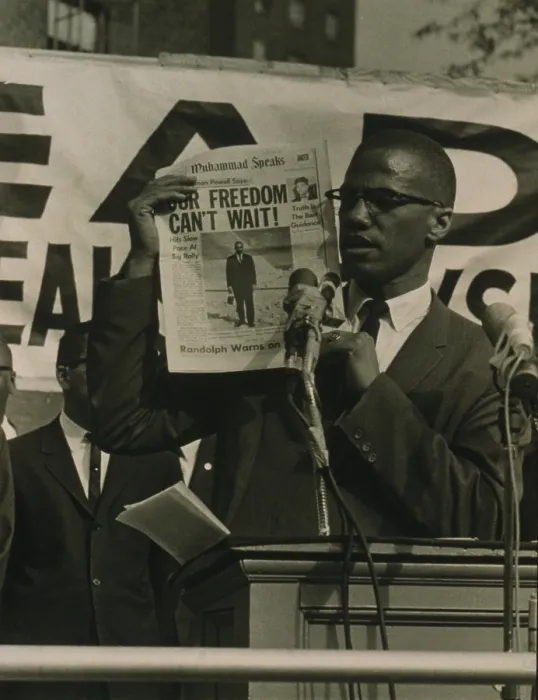 Framed Print : Malcolm X, holding up newspaper with headline Our Freedom Can't Wait, while standing behind podium with microphones addressing a crowd at a Black Muslim rally in New