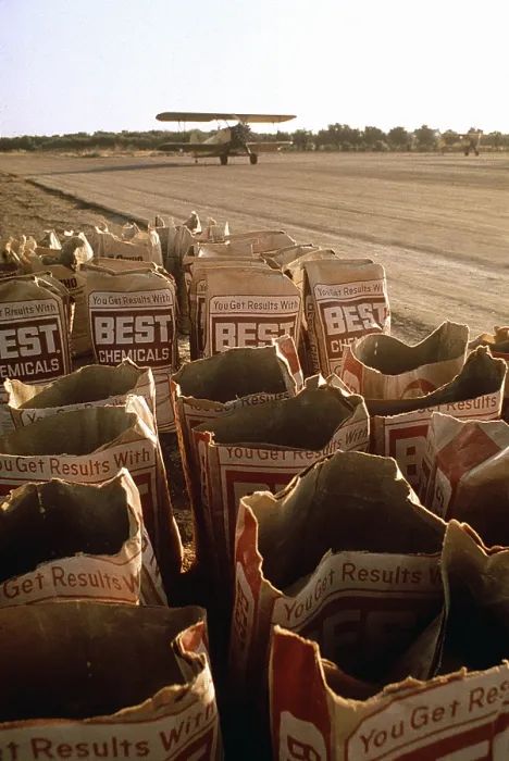 Framed Print : Used bags of Best Chemical bags next to a runway where a bi-plane is about to take off to spread the chemicals over crops near Fresno California. Ca. 1973-75