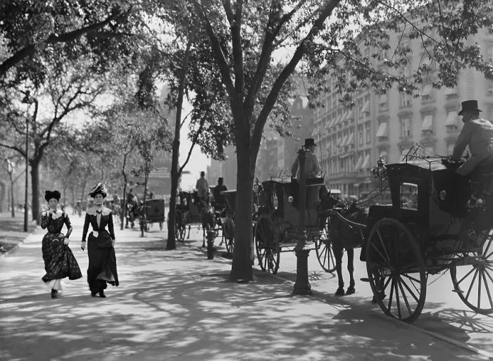 Framed Print : Two young women in fashionable cinched waist dresses, walk on the Madison Avenue sidewalk, NYC. They pass a line of horse drawn cabs, waiting for hire. Photo by Byron, c