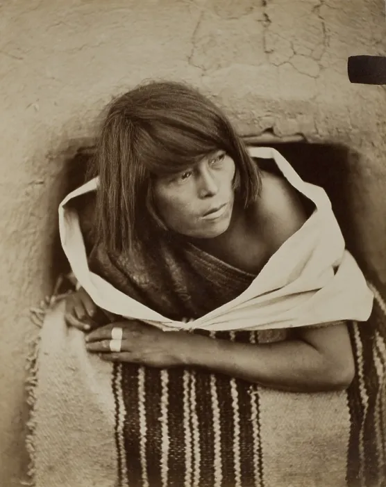 Framed Print : ZUNI, WATCHING, c. 1879-81, by John K. Hillers. A Zuni woman leans out of an adobe window, with her shoulders encircled by loose textile garments