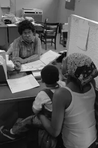 August Prints: An African American woman with a child on her lap talking to a case worker at a New York City welfare office