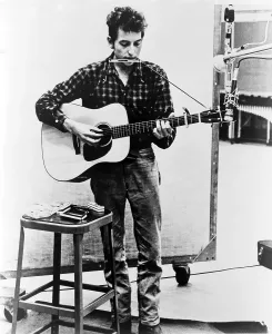 Dylan Prints: Bob Dylan (b. 1941) playing guitar and harmonica into microphone. 1965