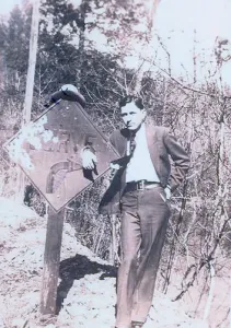 Clyde Prints: Clyde Barrow (1909-1934) dressed in a suit and standing with his hand place through a hold on a road sign