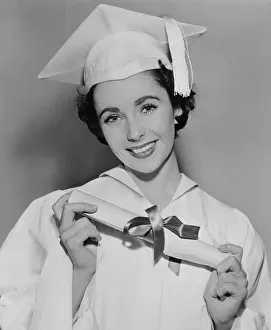 Cap And Gown Prints: Elizabeth Taylor (b. 1932) wearing white cap and gown while holding a diploma from high school in Los Angeles
