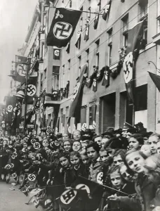 Germany Prints: German children equipped with swastika flags look toward an advancing Nazi parade