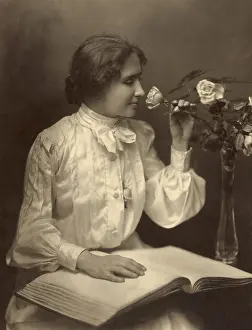 Communication Prints: Helen Keller seated with her hand on braille book as she smells a rose in a vase, 1904