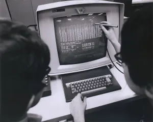 Progress Prints: IBM programmers work at a cathode ray monitor with a touch-screen pointer. Ca. 1980
