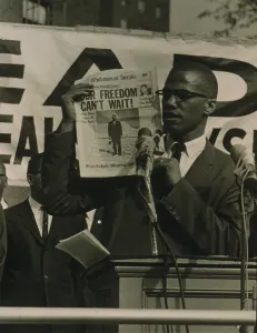 Malcolm X, holding up newspaper with headline Our Freedom Can't Wait, while standing behind podium with microphones addressing a crowd at a Black Muslim rally in New