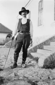 1880s Portaits Prints: Serbian man wearing hat, vest, belted pants and pointed shoes with cane standing outside of building in Yugoslavia