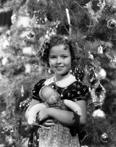 Temple Prints: Shirley Temple holding doll in front of Christmas tree, ca. late 1930s