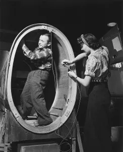 December Prints: A small man installs control wires inside the fuselage of a Valiant basic trainer while a woman works standing outside