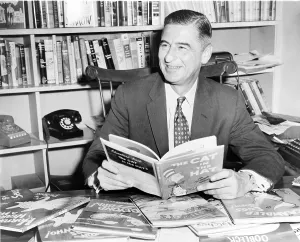 Cartoonist Prints: Ted Geisel (Dr. Seuss) (1904-1991) seated at desk covered with his uniquely humorous children's books